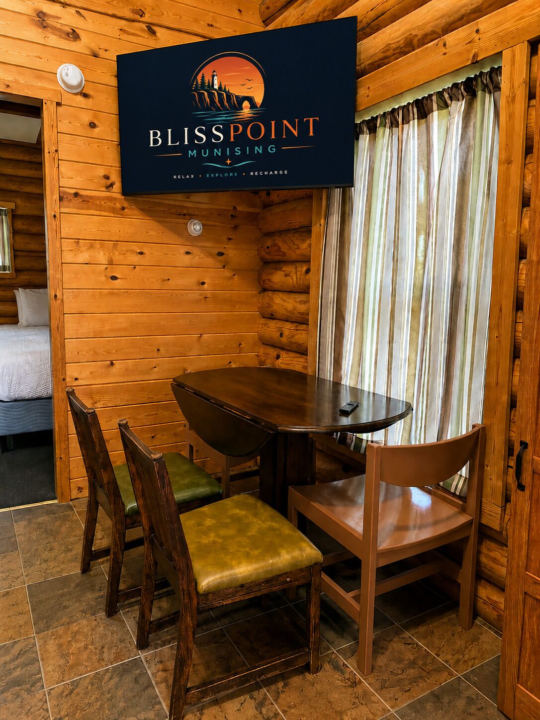 A wooden cabin interior with a round table and chairs, a bed, and a sign reading 'BLISSPOINT MUNISING' with the words 'RELAX,' 'EXPLORE,' and 'RECHARGE.'