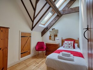 Un dormitorio con techo inclinado, vigas de madera, tragaluces, cama con ropa blanca, una silla roja, cómoda de madera y armario de madera.