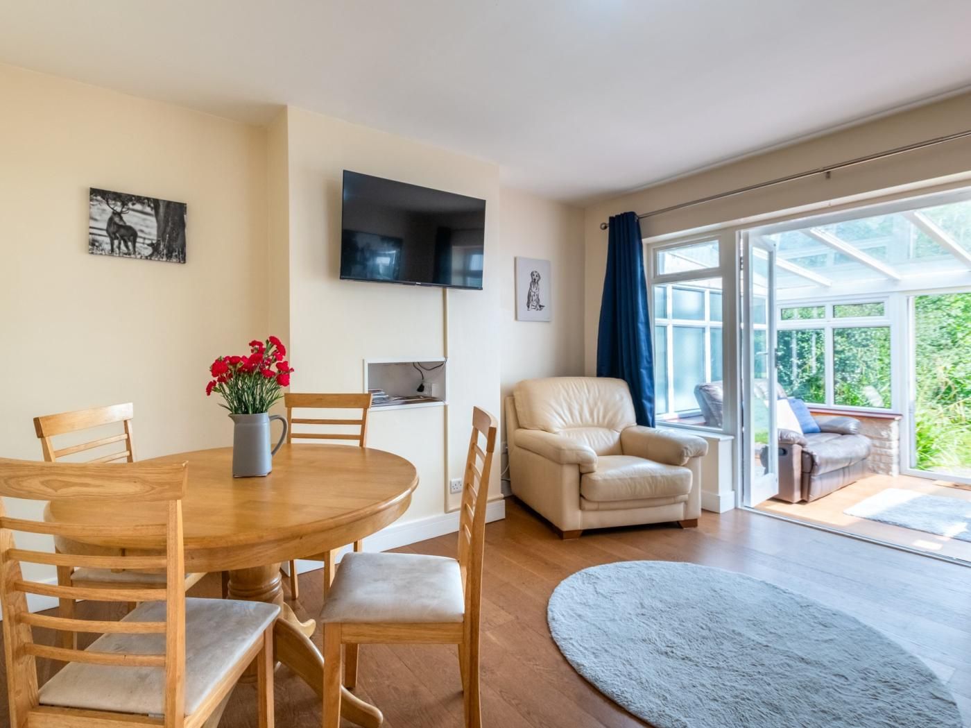 Una habitación con una mesa de comedor de madera, sillas, un sofá, un televisor montado en la pared y un amplio ventanal con cortinas.