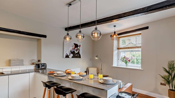 Una cocina moderna con una isla central, taburetes de bar, iluminación colgante y un ventanal con persianas.