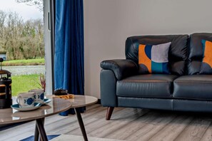 Ein modernes Wohnzimmer mit einer Ledersofa, einem runden Couchtisch und Blick auf einen Garten durch ein Fenster mit blauen Vorhängen.