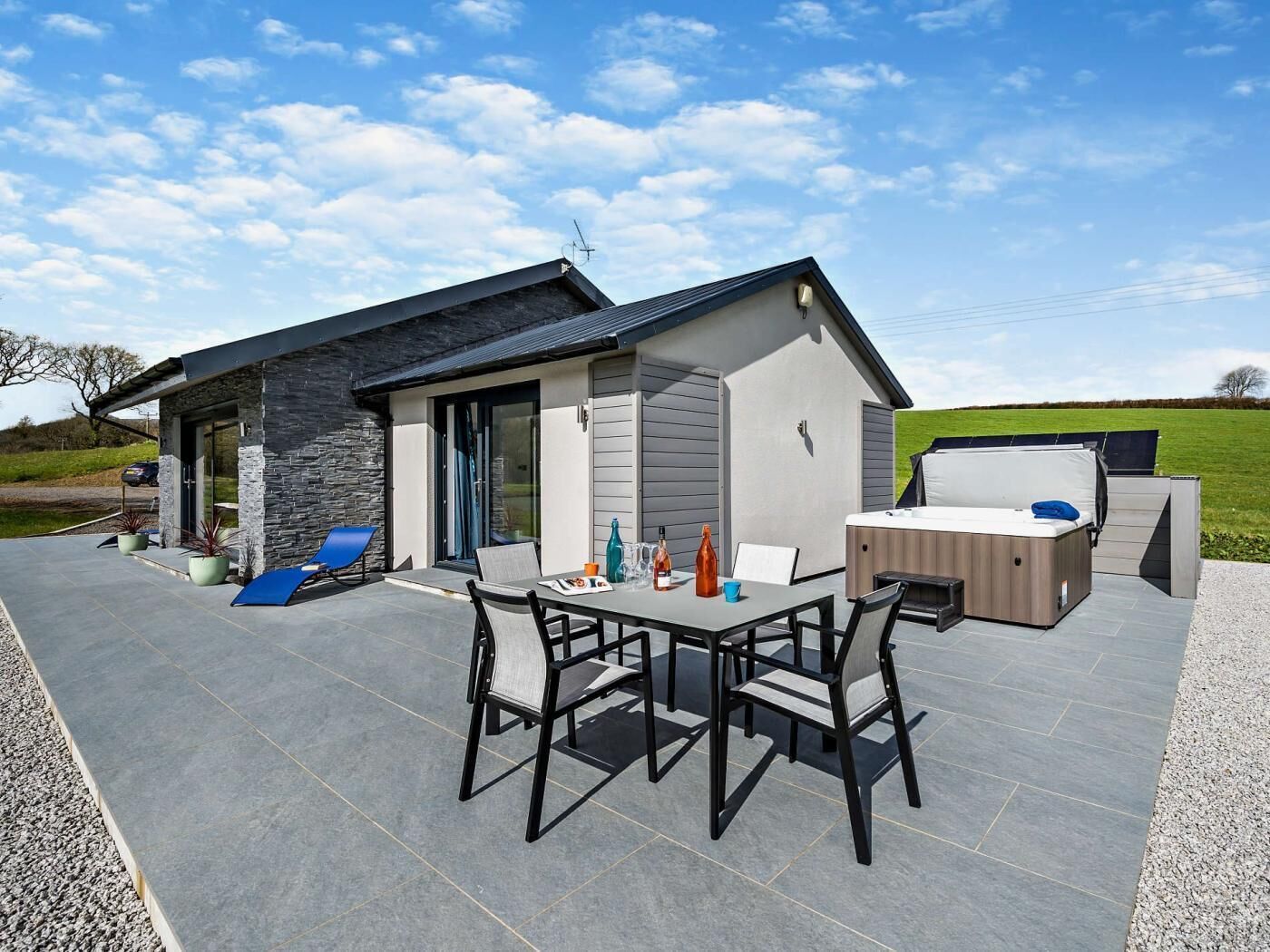 A modern outdoor patio with a dining table and chairs, a hot tub, and a stone wall.