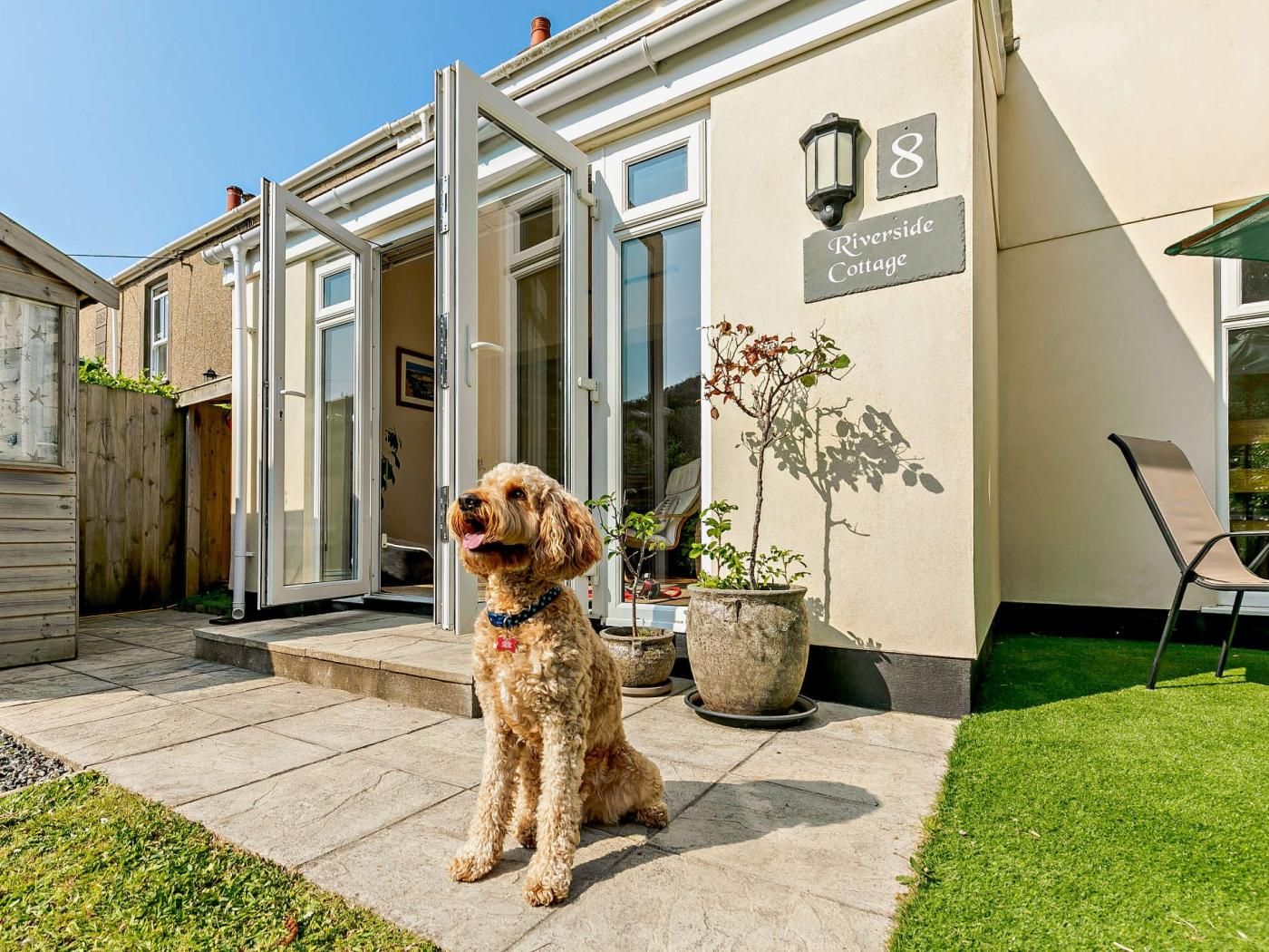 A dog sitting outside a house with a sign reading 'Riverside Cottage.'