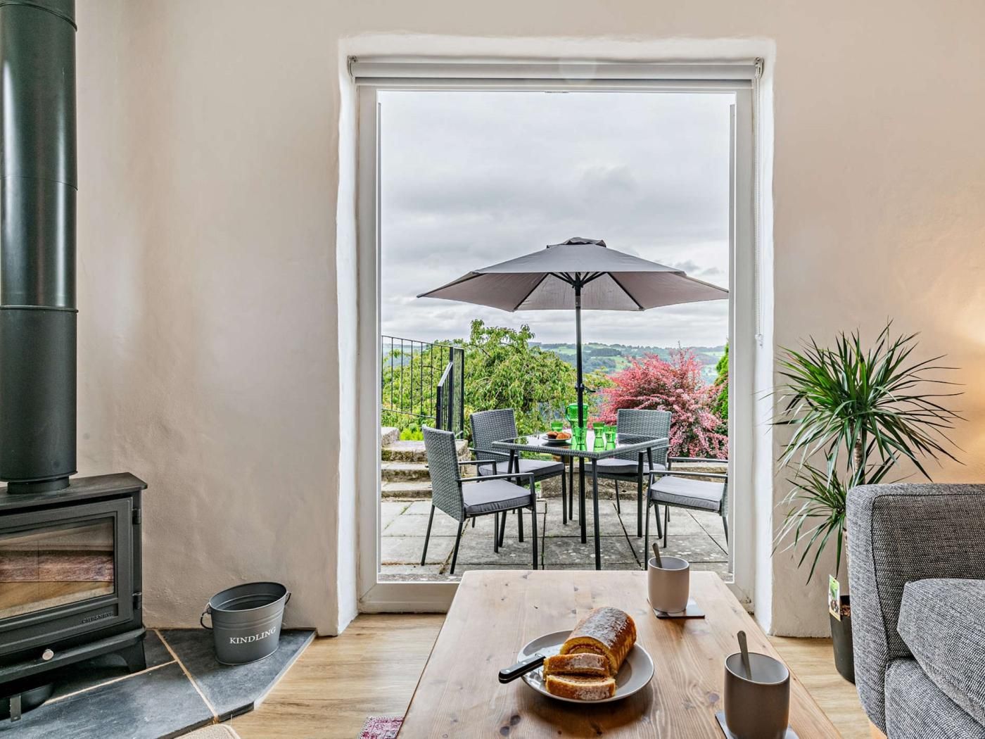 Un espace de vie chaleureux avec un poêle à bois, une table à manger où est posée une miche de pain, et un patio aménagé avec des chaises et un parasol.