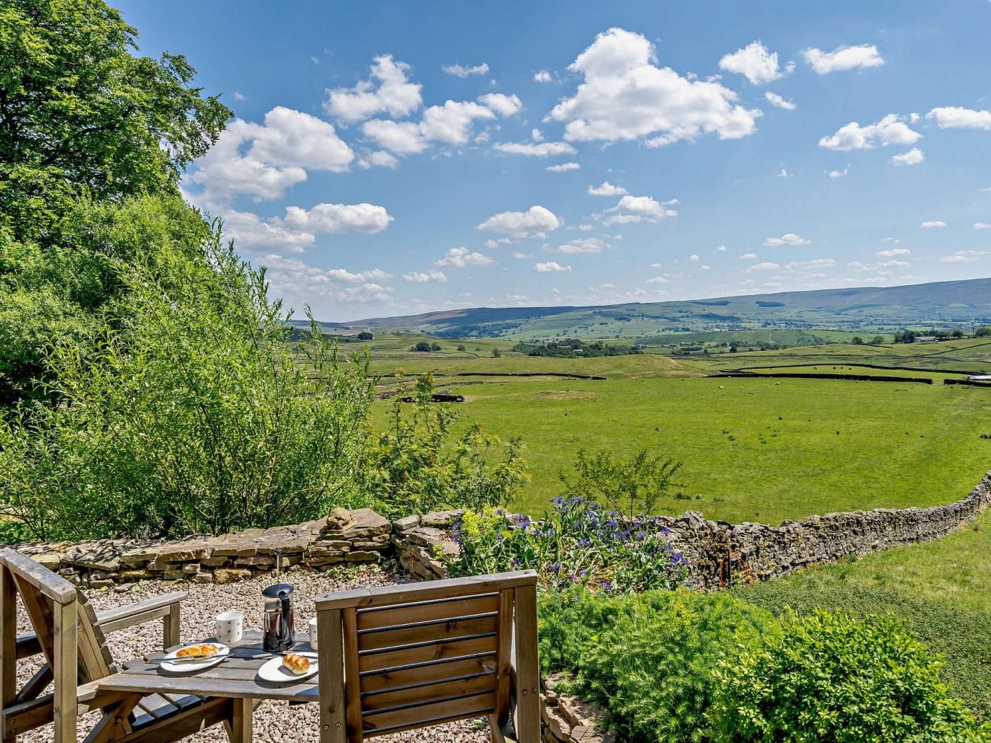 An outdoor dining area with a table and chairs, overlooking a green field and distant hills.