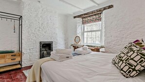 A bedroom with a white brick wall, a wooden dresser, a bed with white bedding, a mirror, a fireplace, and a window with a patterned curtain. - 3 Bed in Fowey (oc-92548) (Fowey)