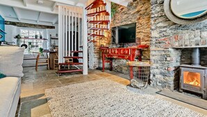 A cozy living room with a fireplace, a TV mounted on a stone wall, a wooden staircase, and a dining area with a view of the outdoors. - 3 Bed in Fowey (oc-92548) (Fowey)