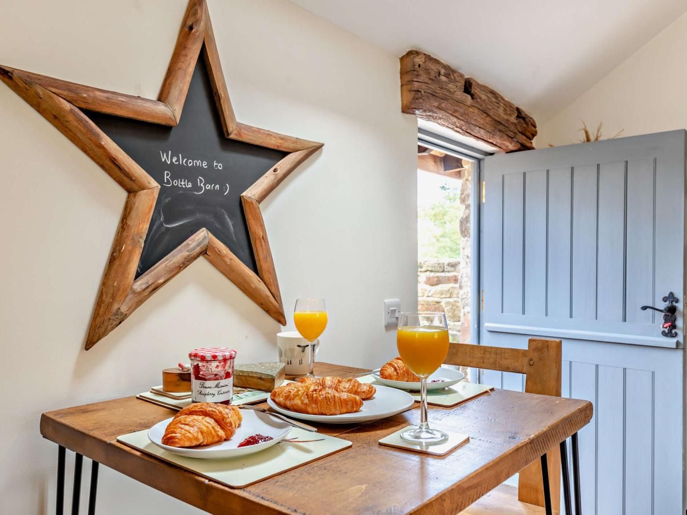 Taula de petit esmorzar amb croissants, un got de suc d'aranja i un botic de melmelada. Un gran rètol de fusta amb forma d'estrella amb la inscripció 'Welcome to Bottega Barn' a la paret.