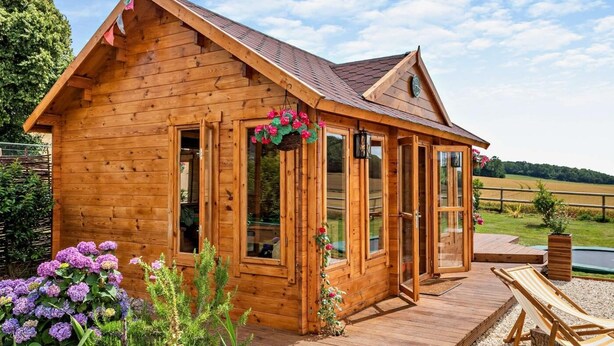 Une cabane en bois dotée de grandes fenêtres, d’une terrasse et d’un jardin fleuri.