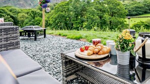An outdoor seating area with a table set for a meal, a wicker chair, and a view of a green landscape. - 1 Bed in Penrith (oc-92641) (Deepdale)