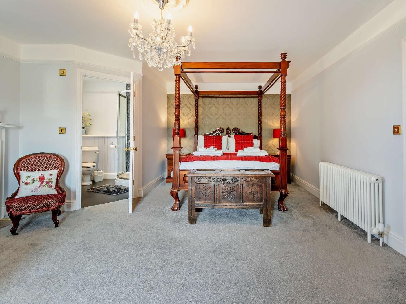 Un lit à baldaquin avec une structure en bois et une tête de lit en marbre, un lustre, un fauteuil recouvert de tissu rouge et un radiateur blanc.