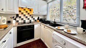 A compact kitchen with white cabinets, a checkered backsplash, a built-in oven, a dishwasher, and a window with blinds. - 2 Bed in Pwllheli (oc-92561) (Llithfaen)