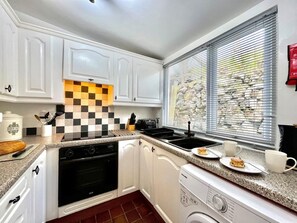 Eine kleine Küche mit weißen Schränken, einer karierten Spritzschutzleiste, einem eingebauten Backofen, einem Geschirrspüler und einem Fenster mit Jalousien.