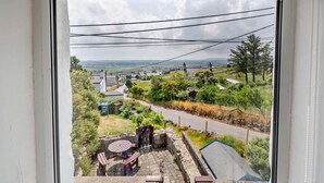 A view through a window of a residential area with a paved driveway, a stone patio with a table and chairs, and a distant landscape with houses and trees. - 2 Bed in Pwllheli (oc-92561) (Llithfaen)