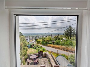 Blick durch ein Fenster auf eine Wohngegend mit befestigter Auffahrt, einer Steinterrasse mit Tisch und Stühlen sowie einer entfernten Landschaft mit Häusern und Bäumen.