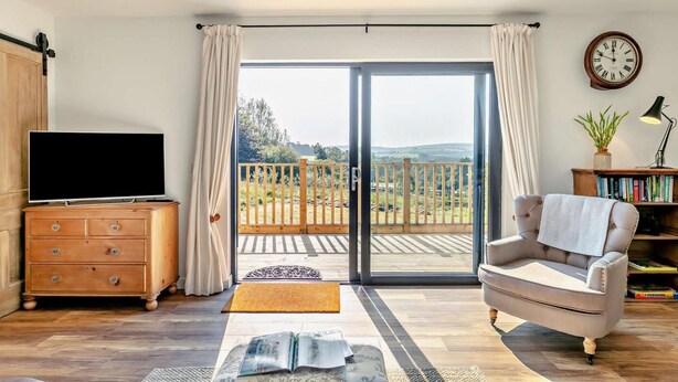 A living room with a TV, a sofa, a bookshelf, and a view of a balcony with a wooden railing. - 2 Bed in Barnstaple (oc-92653) (Barnstaple)