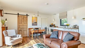A modern living room with a leather sofa, a dining table, and a kitchen area with a window. - 2 Bed in Barnstaple (oc-92653) (Barnstaple)