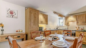 Une table à manger en bois, avec des chaises, est dressée pour six personnes. On aperçoit une cuisine au fond et un tableau encadré est accroché au mur.