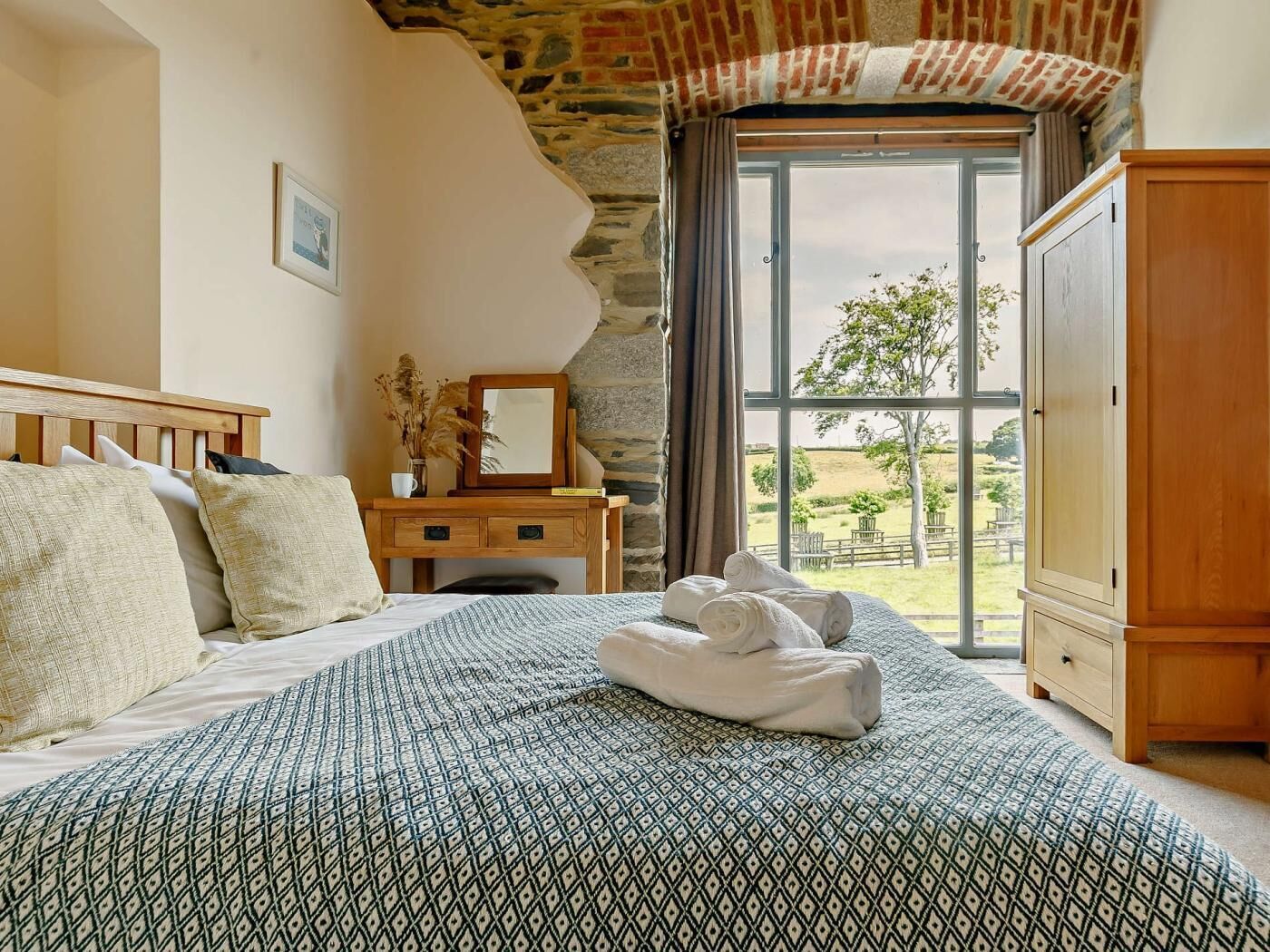 Een slaapkamer met een bed met een patroon, een houten bureau met een spiegel en uitzicht op het landschap door een groot raam.