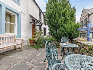 A courtyard with stone flooring, a wooden bench, metal chairs, a table with cups, and a large coniferous tree. - 2 Bed in Keswick (oc-92600) (Keswick)