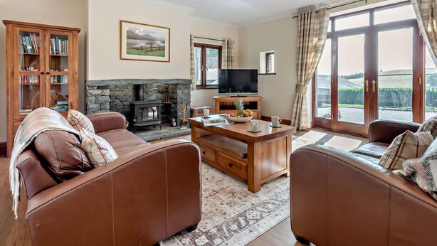 A cozy living room with a stone fireplace, leather sofas, a television, and a large window with curtains. - 3 Bed in Wigton (oc-92500) (Uldale)