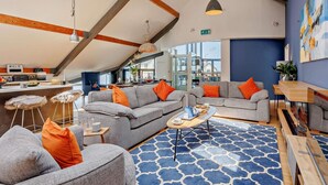 Un interno moderno con soffitto a mansarda, divani grigi con cuscini arancioni, un tappeto azzurro decorato e un tavolo in legno con tazza e piattino.