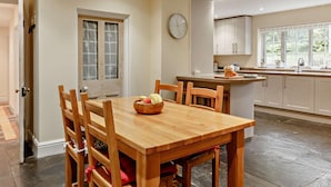 A dining area with a wooden table and chairs, a kitchen with white cabinets, a clock on the wall, and a window with a view of greenery. - 4 Bed in Tavistock (oc-92491) (Gulworthy)