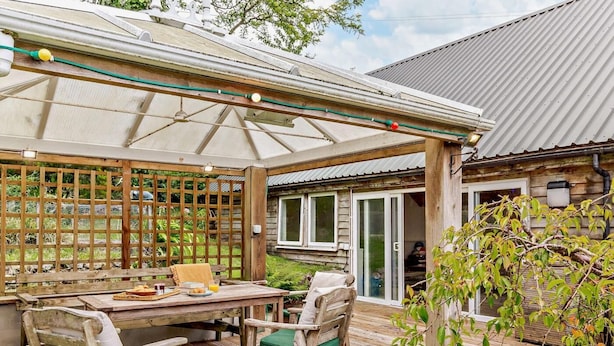 A wooden deck with a table and chairs, a pergola with lights, and a view of a house with a sliding door. - 4 Bed in Tavistock (oc-92491) (Gulworthy)