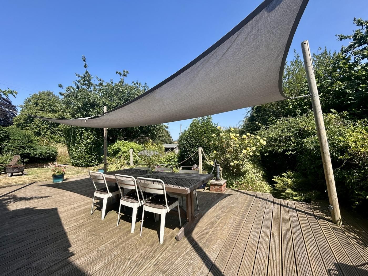 A wooden deck with a table and chairs, surrounded by greenery and a large shade sail.