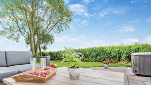 Una terrazza esterna con tavolo in legno, divano grigio, vasca idromassaggio e siepe sullo sfondo.