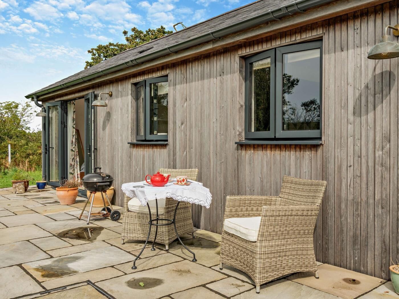 A wooden building with a patio area, a table with a white tablecloth, a wicker chair, and a barbecue grill.