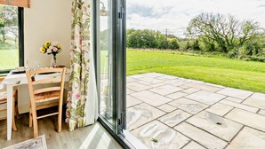 Ein Zimmer mit Esstisch, Stühlen, einer Blumenvase und einer Schiebetür zu einer Terrasse mit Rasen und Bäumen.