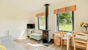 Ein gemütliches Wohnzimmer mit Kamin, Fernseher, einer Couch, einem Esstisch mit Stühlen und großen Fenstern mit Blick auf Grünflächen.
