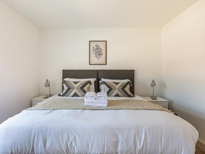 Ein ordentlich bezogenes Bett mit weißer Decke und zwei Kissen, ein gerahmtes Kunstwerk darüber sowie je eine Nachttischlampe auf beiden Seiten.