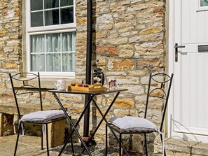 A stone building exterior with a white door, a window with curtains, and a table with chairs and a tray of items. - 2 Bed in Consett (oc-92807) (Hunstanworth)