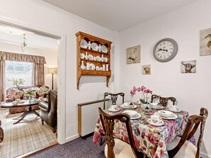 A dining area with a floral tablecloth, wooden chairs, a wall-mounted clock, and a shelf with decorative plates. - 2 Bed in Consett (oc-92807) (Hunstanworth)