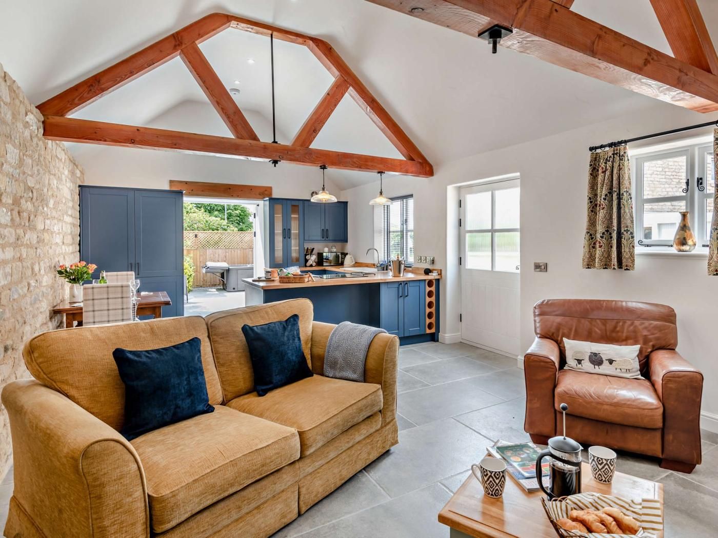 A spacious living area with a sofa, a coffee table, and a leather armchair. A kitchen with blue cabinets and a dining area with a table and chairs are visible in the background.