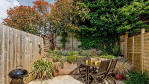 Ein Garten mit Holzzaun, Grill, Tisch mit Stühlen und verschiedenen Kübelpflanzen.