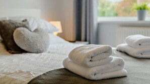 Ein ordentlich bezogenes Bett mit weißer Decke, einem herzförmigen Kopfkissen und einem Stapel gefalteter Handtücher.