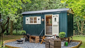 En liten, grön byggnad med ett tak av metall, en träveranda, ett bord, stolar och en liten trädgårdsdel.