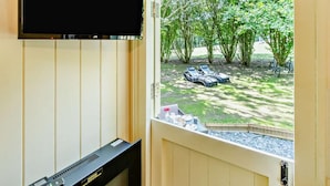 Ett rum med en tv som är monterad på väggen, ett piano och en dörr som leder ut till ett utomhusområde med solstolar och träd.