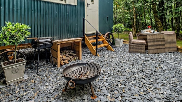 En rustik utomhusmiljö med en grill av trä, ett litet bord med stolar och en byggnad med vågplåtsfasad.