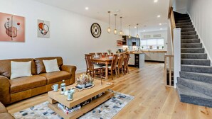 Una sala de estar moderna con un sofá de cuero marrón, una mesa de centro de madera y un comedor con sillas de madera, además de una cocina al fondo.