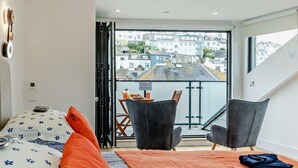 Una habitación moderna con cama, dos sillas, una mesa con productos de desayuno y un balcón con vista a la ciudad.