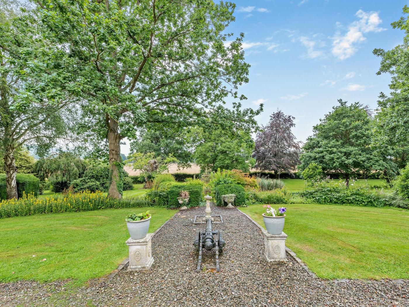 A garden pathway with a central water feature, surrounded by lush greenery and trees.