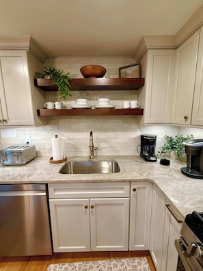 A kitchen with white cabinets, a stainless steel dishwasher, a sink, a toaster, a coffee maker, and a wooden shelf with dishes and a plant. - Newly renovated 2bed/2 bath, pet friendly, easy walk to village! (Skaneateles)