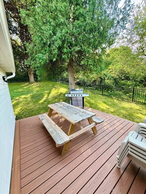 A wooden deck with a picnic table and grill, surrounded by lush greenery and a black metal fence. - Newly renovated 2bed/2 bath, pet friendly, easy walk to village! (Skaneateles)