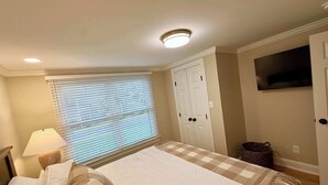 Ein Schlafzimmer mit einem Bett, einem Fenster mit Jalousien, einer Deckenleuchte, einem Kleiderschrank und einem an der Wand befestigten Fernseher.