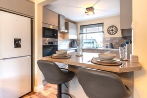 A modern kitchen with a built-in oven, microwave, and water dispenser. There is a dining area with a table set for two, and a large window providing natural light. - 2 Bed in Seahouses (oc-92880) (Seahouses)
