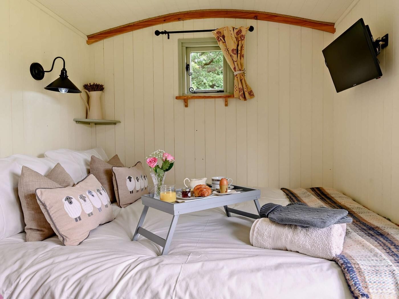 A cozy bedroom with a bed, pillows, a bedside table with a tray of breakfast, a wall-mounted TV, and a window with curtains.